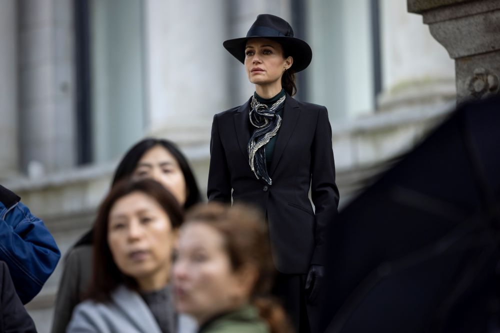 Verna wears a black suit with a black fedora while standing outside in a crowd in front of a courthouse in The Fall of the House of Usher Season 1 Episode 8, "The Raven."