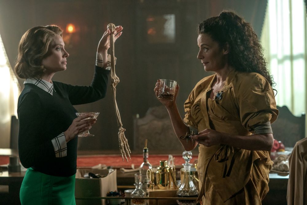 Rita holds up a skeleton arm in front of Laura, who holds a glass of liquor, while standing in the dining room in Doom Patrol Season 4 Episode 7, "Orqwith Patrol."