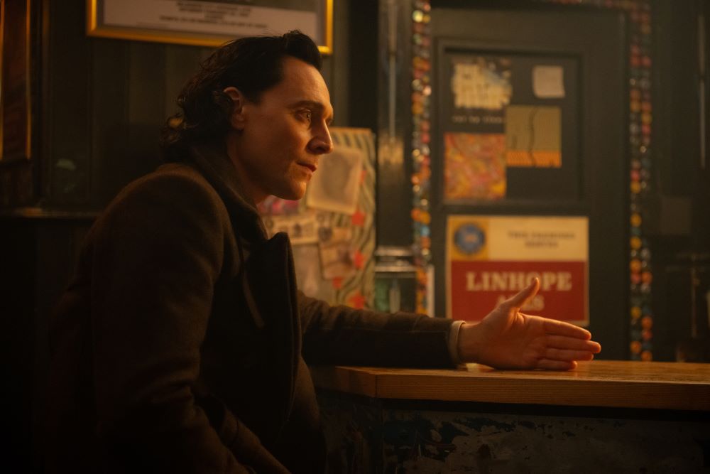 Loki sits at a bar in a dimly lit pub while gesturing in Loki Season 2 Episode 5, "Science/Fiction."