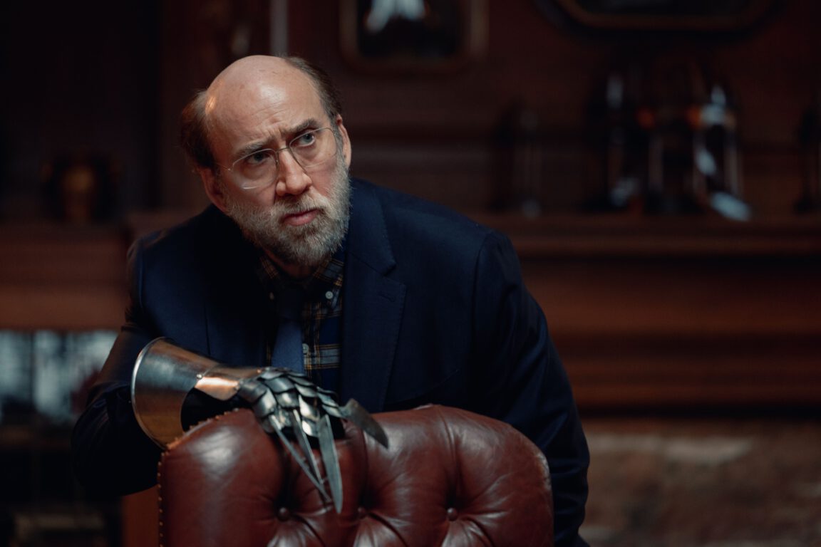 Nicolas Cage looks pensive as he wears a medieval gauntlet on his hand in the movie Dream Scenario. 