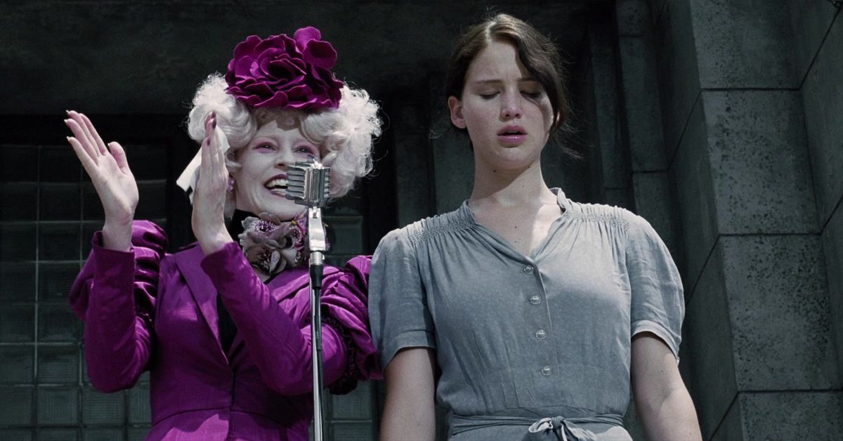 Katniss, a young white woman in a simple blue dress stands at a podium with Effie, a woman in a blonde wig and maroon fascinator, and maroon skirt suit.