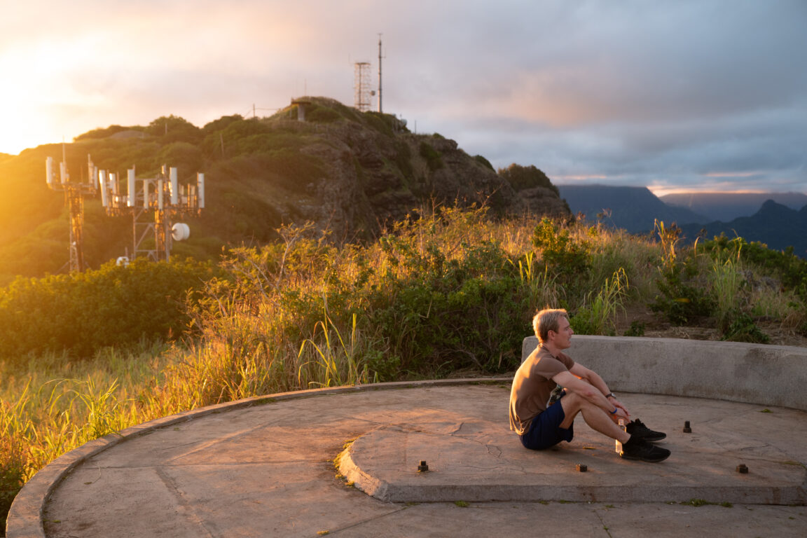 Michael Fassbender sits on a hillside during sunset appearing deep in thought in the movie Next Goal Wins. 