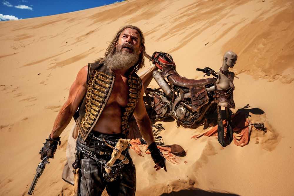 Chris Hemsworth wears a leather vest and pants with no shirt while sporting long hair and a beard as he stands in the desert for "Furiosa: A Mad Max Saga."