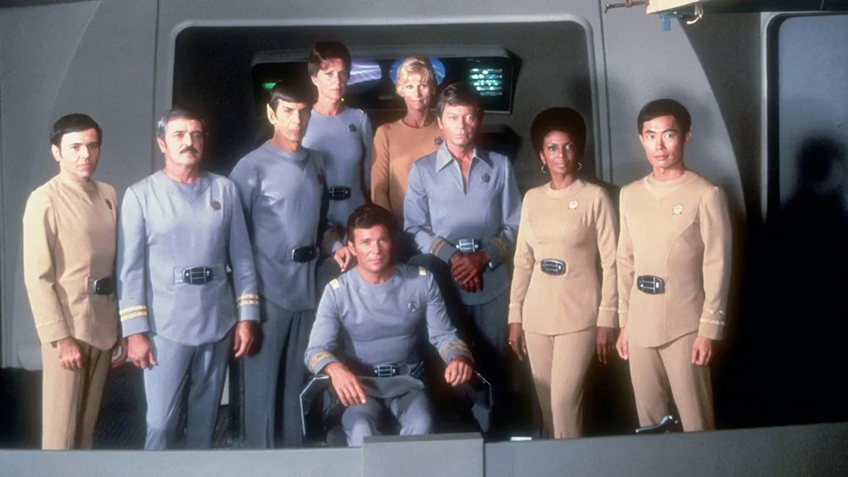 Chekov (Walter Koenig), Scotty (James Doohan), Spock (Leonard Nimoy), Chapel (Majel Barrett Roddenberry), Rand (Grace Lee Whitney), Kirk (William Shatner),McCoy (DeForest Kelley), Uhura (Nichelle Nichols) and Sulu (George Takei). They are all in the TMP Starfleet uniforms and posing on the Enterprise bridge.