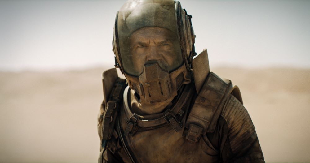 Gurney Halleck wears a helmet and a space-type suit while standing outside in the desert.