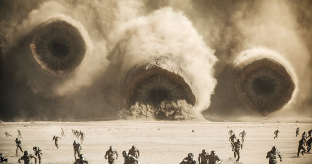 A cluster of giant sandworms move through the desert while a group of people flee from them.