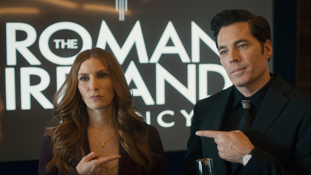 Susan Ireland and Luke Roman stand in front of the logo for The Roman Ireland Agency while pointing at each other in SurrealEstate Season 2 Episode 10, "Letting Go."