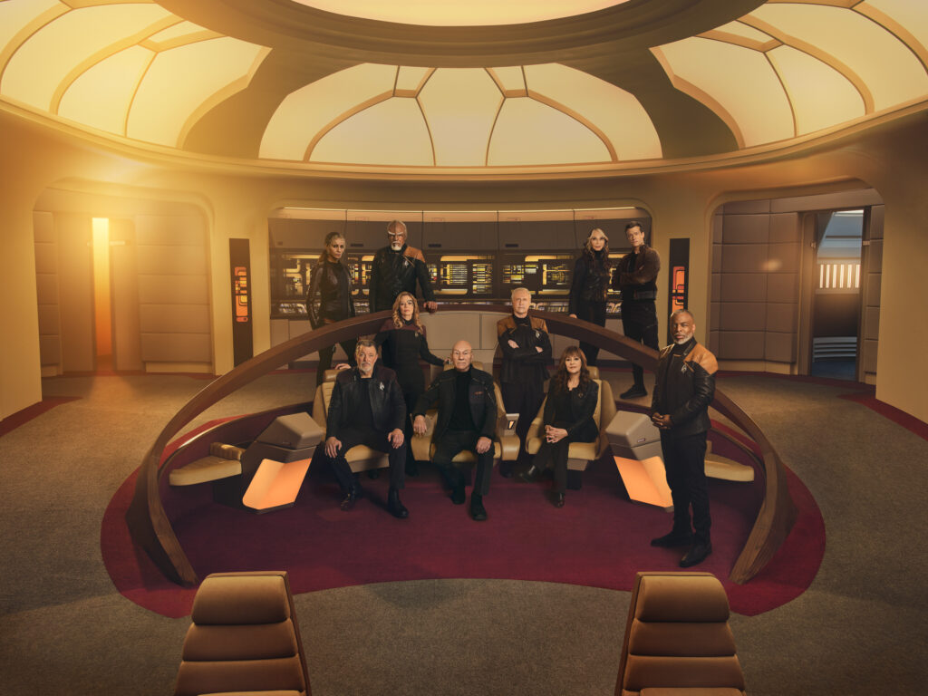 Jonathan Frakes as Riker, Jeri Ryan as Seven of Nine, Sir Patrick Stewart as Picard, Brent Spiner as Data, LeVar Burton as Geordi, Michelle Hurd as Raffi, Michael Dorn as Worf, Gates McFadden as Beverly Crusher and Ed Speleers as Jack Crusher in Star Trek: Picard season 3. They are all posing on the bridge of the Enterprise-D.