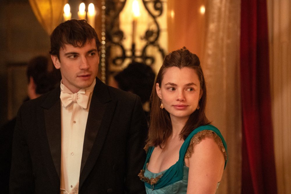 Theo and Nan wear Victorian finery while standing in a brightly lit mansion during a party in The Buccaneers Season 1 Episode 7, "First Footing."
