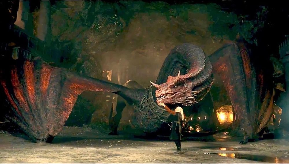 Large, red tinted dragon hunched over a white haired human in a cavernous structure in the series House of the Dragon. 