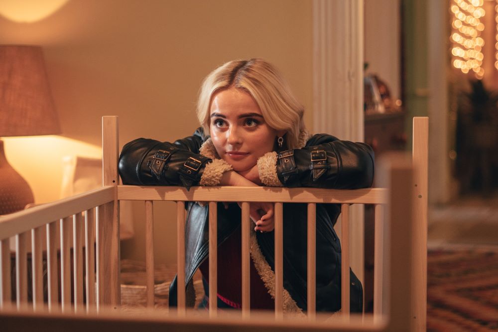 Millie Gibson as Ruby Sunday. She wears a black leather jacket with fleece lining while leaning on a wooden banister in a house in Doctor Who.