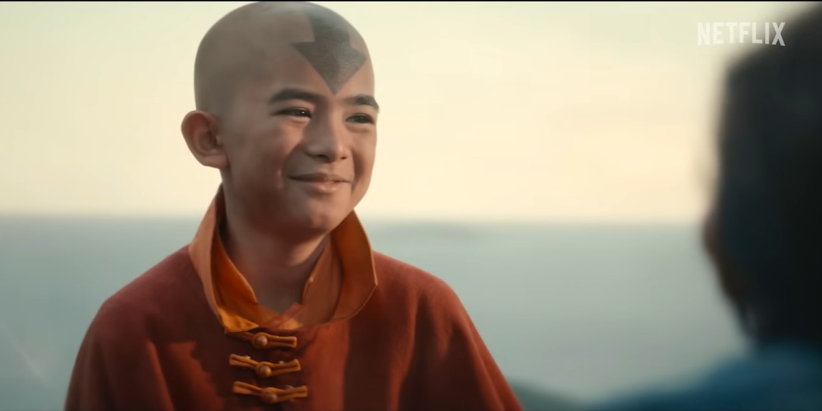 A scene from Avatar: The Last Airbender. Aang, an Asian boy with a bald head and a blue arrow tattoo, smiles at someone offscreen.