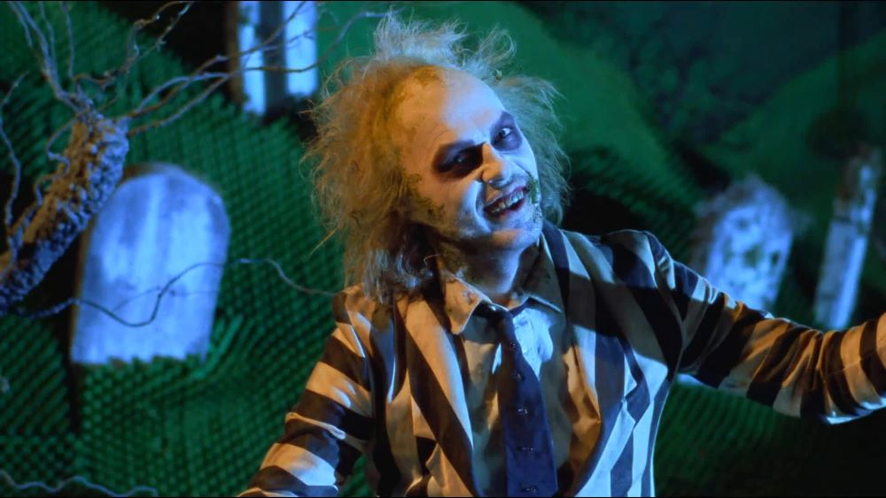 A still of Michael Keaton as Beetlejuice in the 1988 film of the same name. He wears a black and white striped suit with grime on his face. He smiles, sitting in a miniature cemetery.