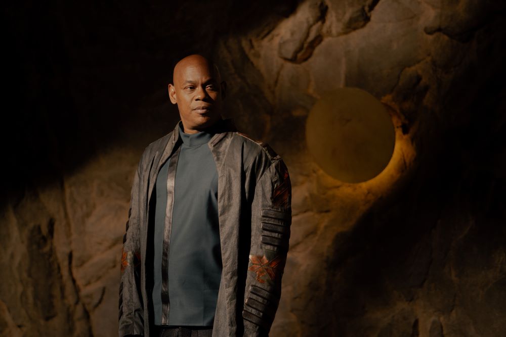 Soren wears a brown leather jacket and black undershirt while standing in his home on an asteroid in Halo Season 2.