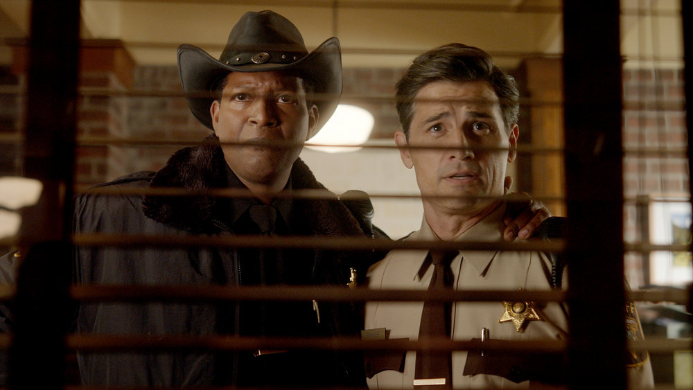 Sheriff Mike wears his black coat with a matching Stetson. He has his arm around Joseph, who wears a deputy uniform. Both stare through window blinds in an office in Resident Alien Season 3 Episode 1, "Lone Wolf."