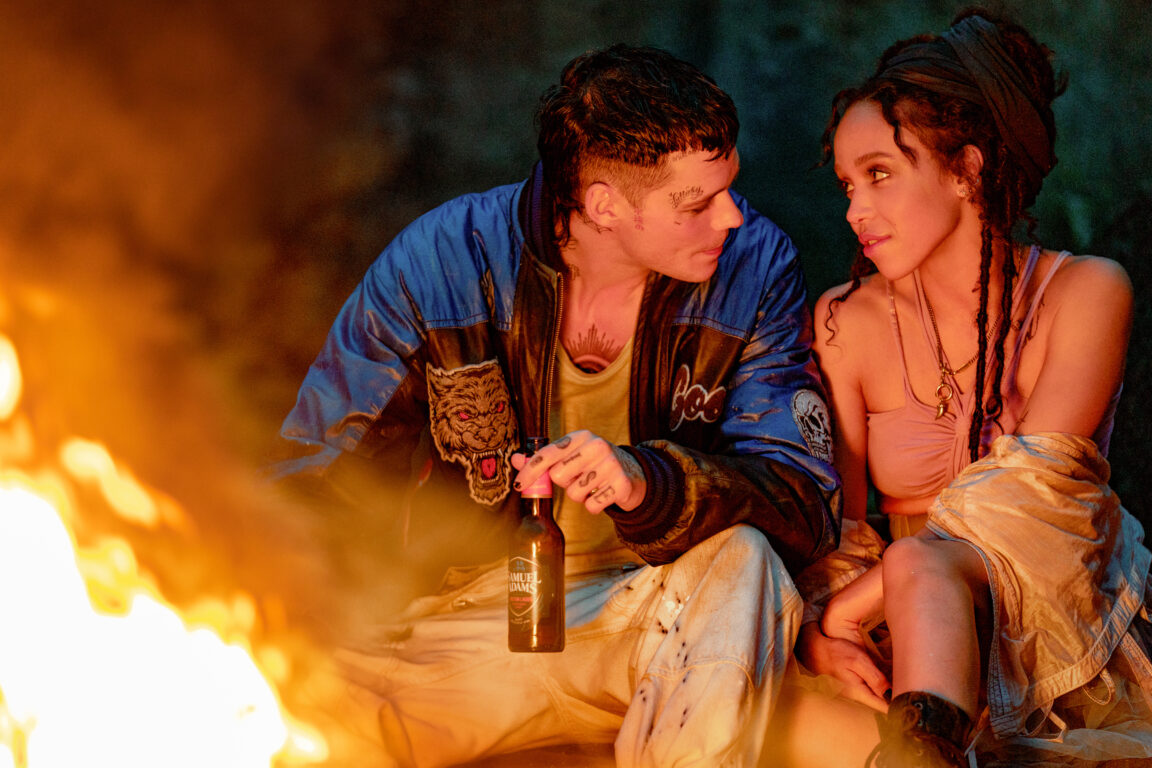 Bill Skarsgård as Eric Draven leans toward FKA twigs as Shelly Webster in a still from The Crow. They're sitting outside in front of a fire and Skarsgård is holding a bottle.