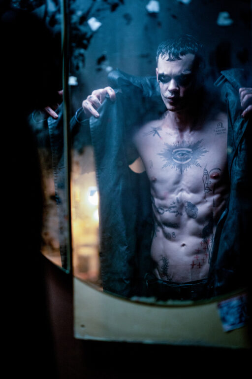 Bill Skarsgård as Eric Draven in a still from The Crow. He's shirtless and putting on a black leather coat while looking in a dirty mirror. His torso is covered in tattoos.