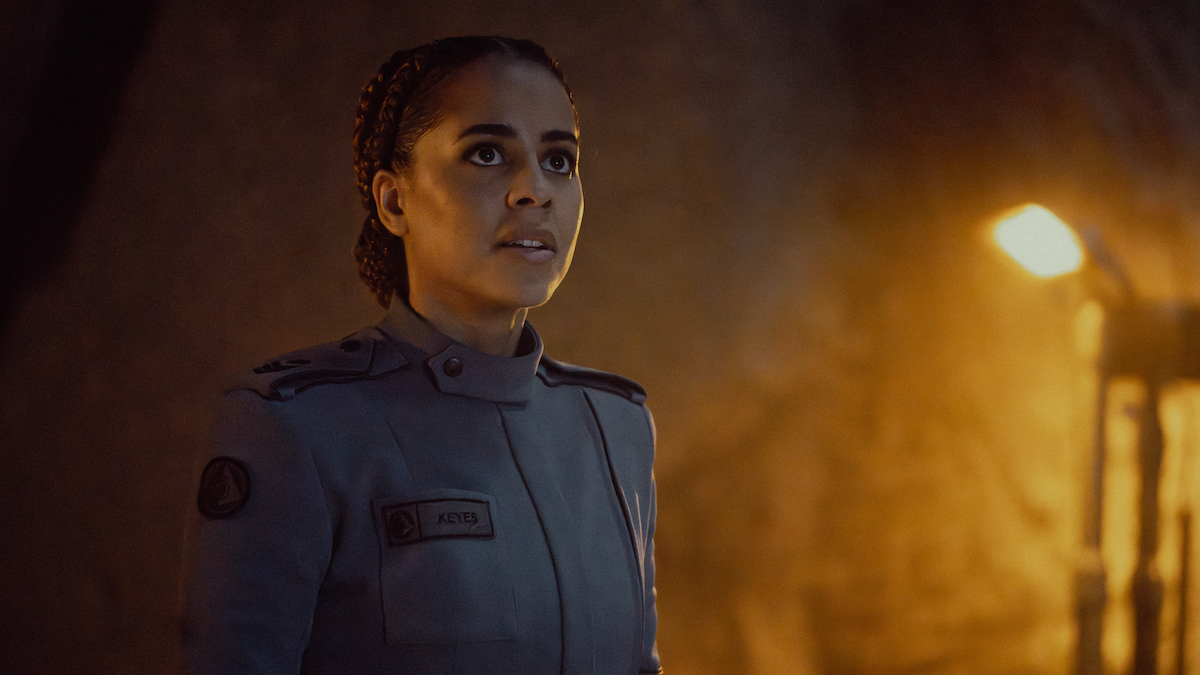 Miranda stands on the left, looking up, dressed in a grey UNSC Science uniform. The background cave walls is lit in golden torchlight in HALO Season 2 Episode 7, "Thermopylae."