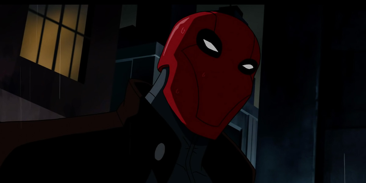 Jason Todd stands in the rain in the city at night. He wears a red mask with a black coat. 