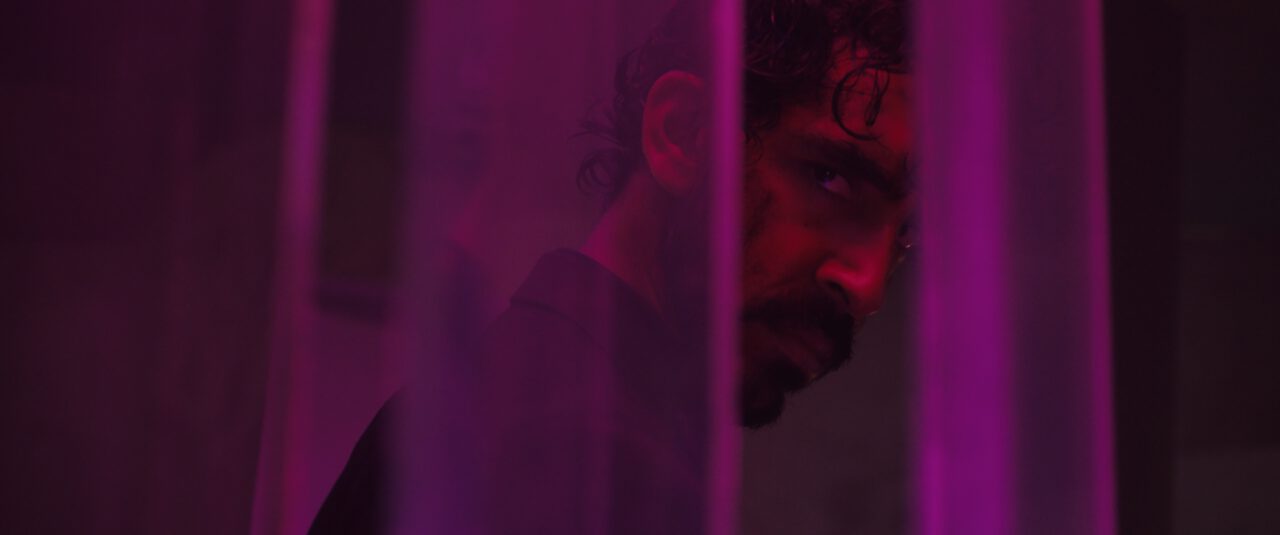 Dev Patel looks out from behind a barrier as he's bathed in purple light in Monkey Man. 