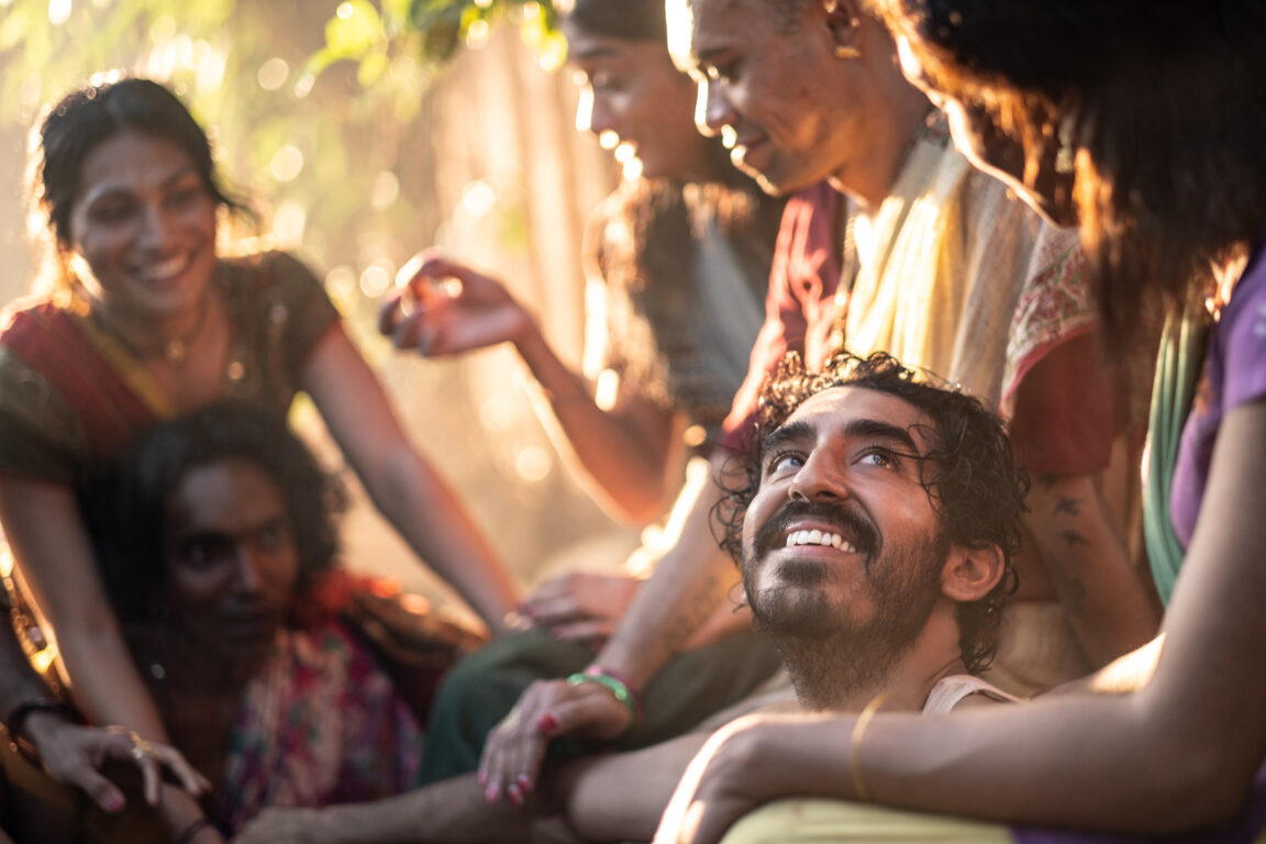 Dev Patel sits in the middle of a group and looks up with a smile on his face in Monkey Man. 