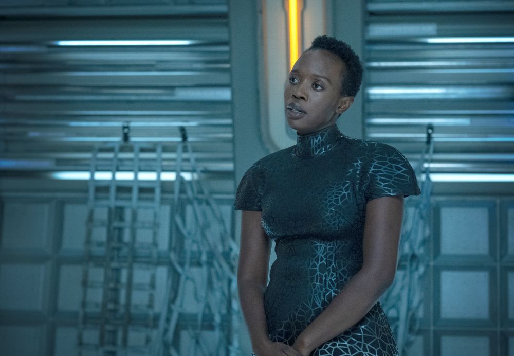 Harmony wears a black leather short-sleeved dress with a silver geometric pattern. She stands in a dimly lit, bluish-tinted room aboard a lighthouse in space on Beacon 23 Season 2 Episode 2, "Purgatory."