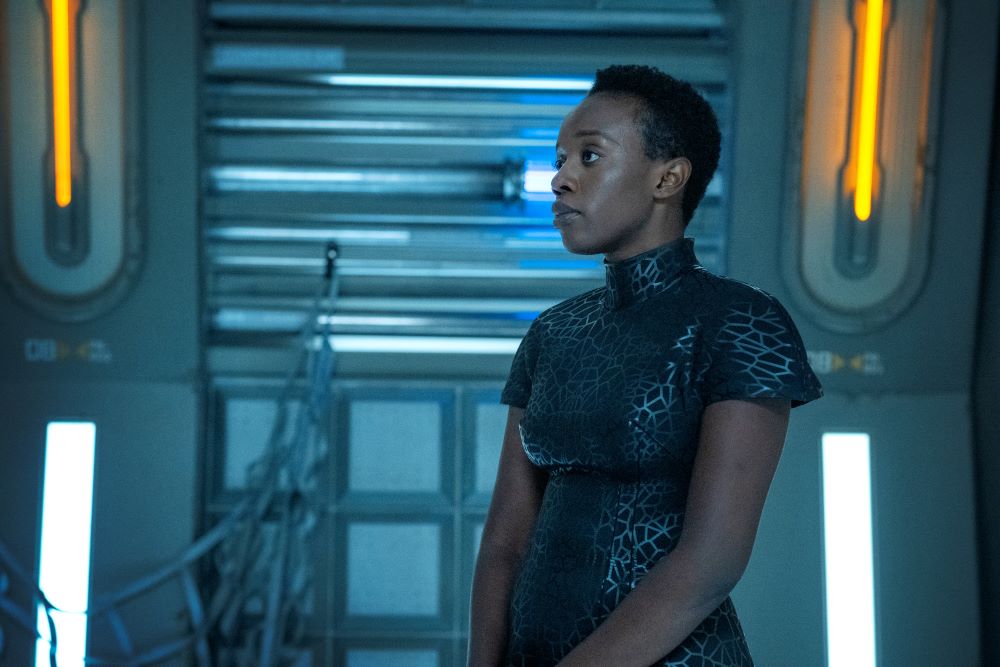 Harmony wears a black leather short-sleeved dress with a silver geometric pattern. She stands in a dimly lit, bluish-tinted room aboard a lighthouse in space on Beacon 23 Season 2 Episode 2, "Purgatory."