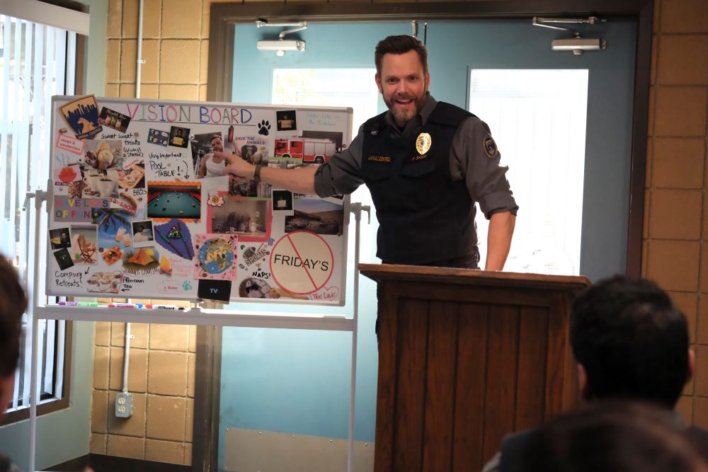 Joel McHale as Frank in the TV comedy Animal Control. He's dressed in an animal control uniform while standing in front of a presentation board while smiling and pointing.