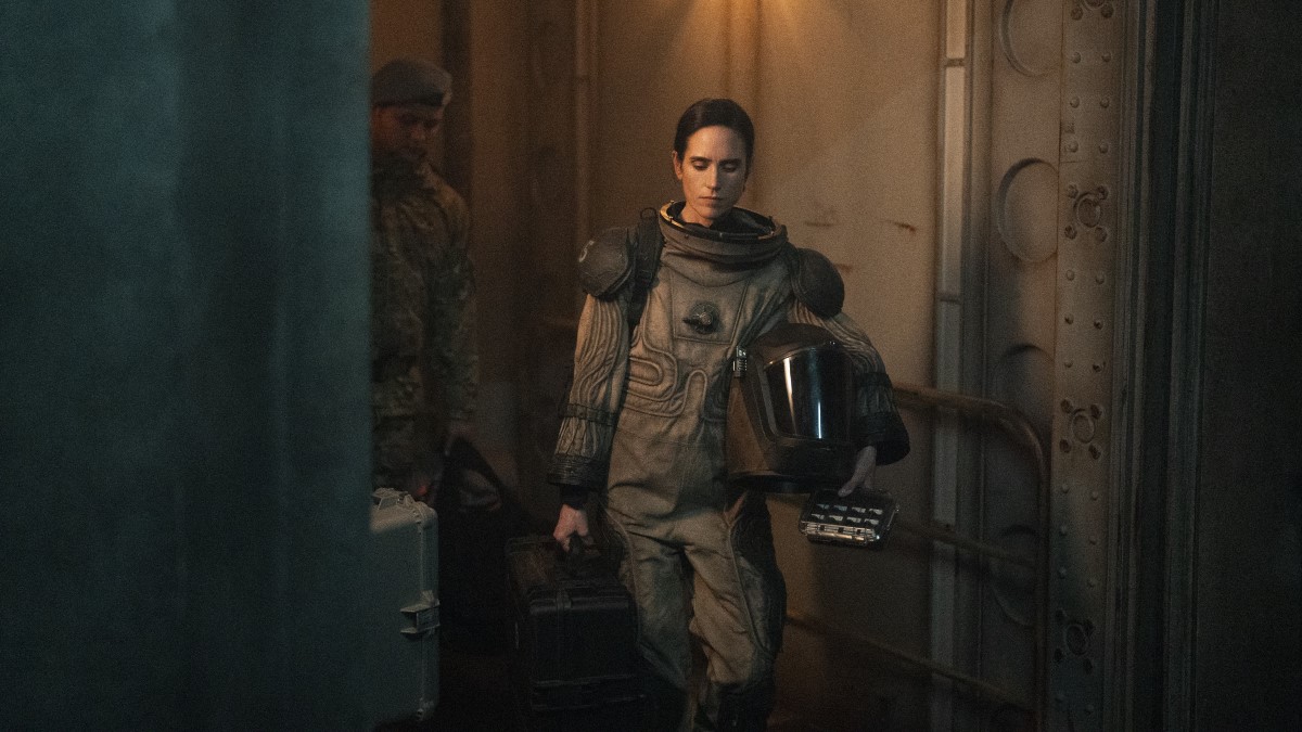 Melanie enters a train car dressed in a beige and grey environmental suit, carrying her helmet under her left arm and a large black case in her right. Snowpiercer Season 4 Episode 7 A Moth to a Flame