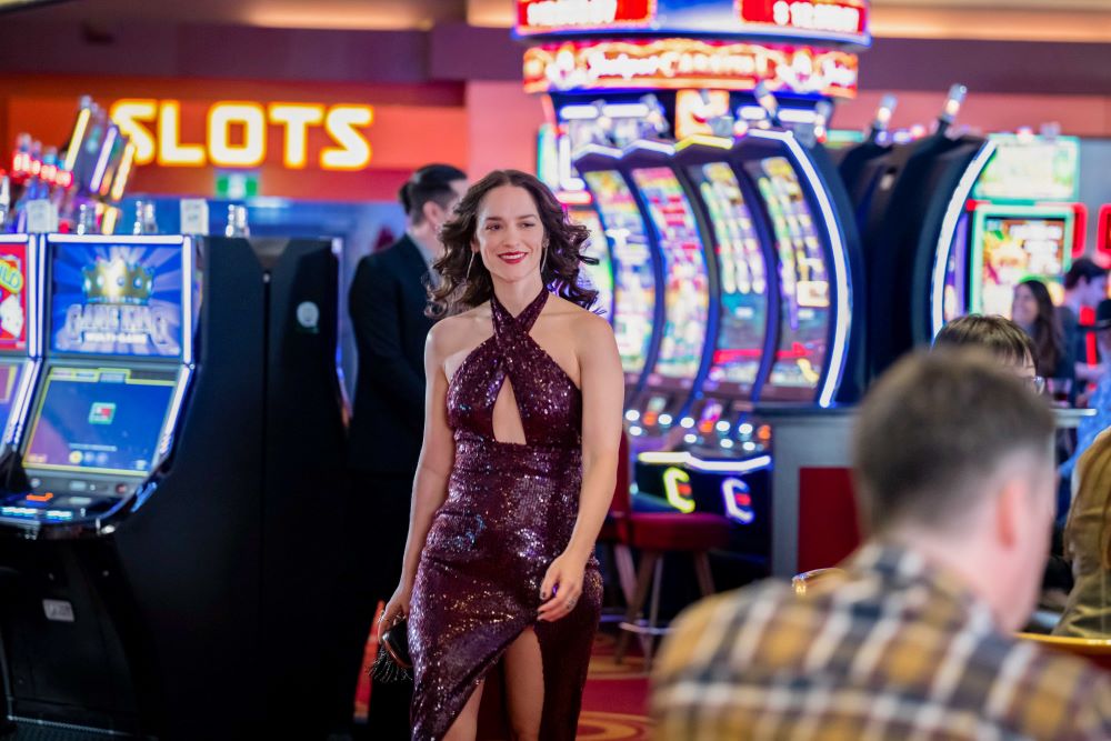 Wynonna Earp wears a sparkling purple halter dress while walking through a busy casino. She smiles.