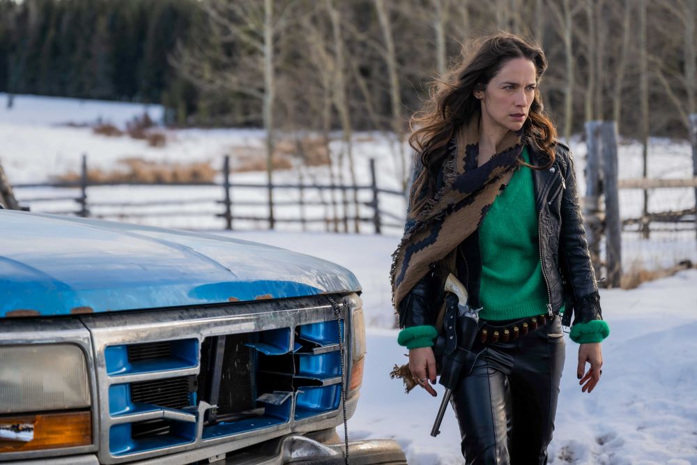 Wynonna holds her gun Peacemaker in her hand as she walks in front of her blue and white pickup truck. She stands on her family property on a snowy day.