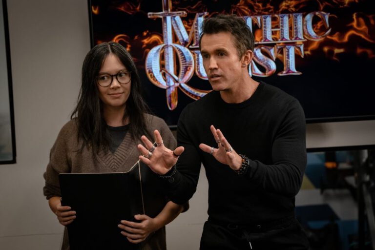Poppy and Ian look focused as they pitch an idea. Poppy holds a folder while Ian gestures and leans forward in Mythic Quest Season 4 Episode 1, "Boundaries."