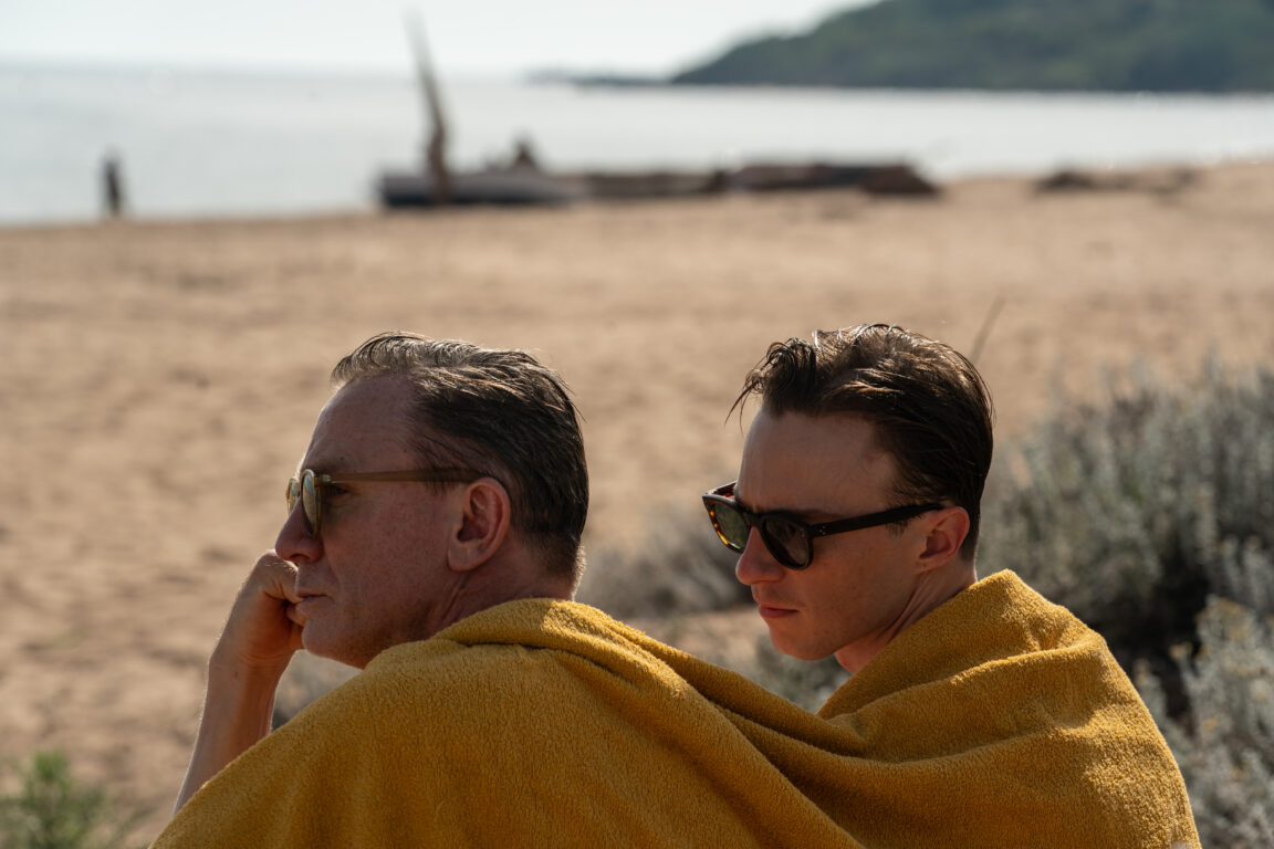 Two men sit on a beach wrapped in towels. They wear sunglasses as they stare out over the water in the movie Queer.