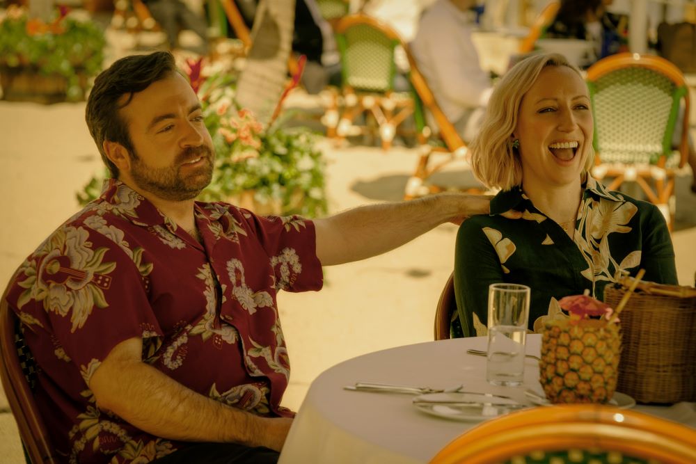 A middle-aged white man and a white woman with blonde hair wear tropical shirts while sitting on the outdoor patio of a restaurant on a sunny day. They're laughing on Side Quest Season 1 Episode 1, "Song and Dance."
