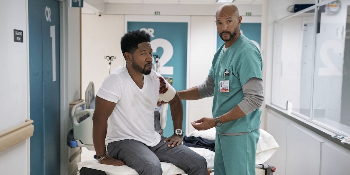 Dante sitting on a gurney while Miles bandages his arm.