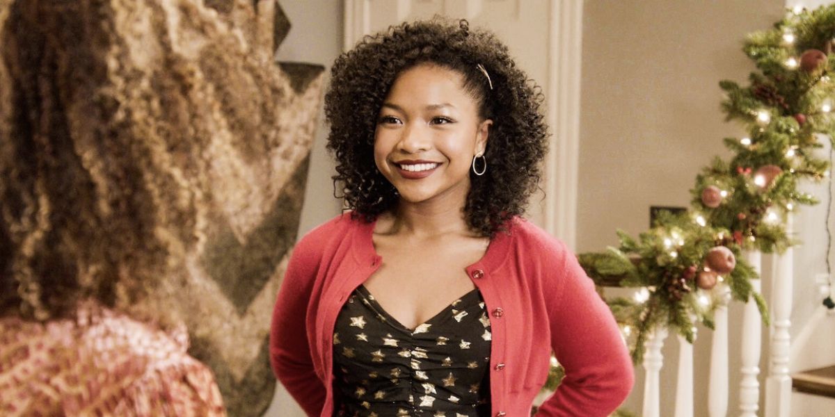 Delilah standing in front of the stairs while smiling and wearing a black dress with stars and a red sweater on The Equalizer.