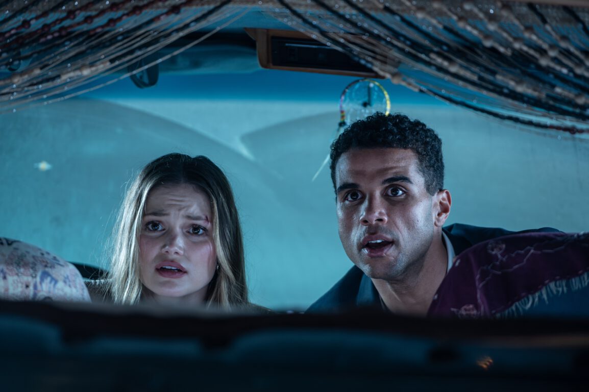 Olivia Holt and Mason Gooding look up over a car seat in surprise in the slasher movie Heart Eyes. 