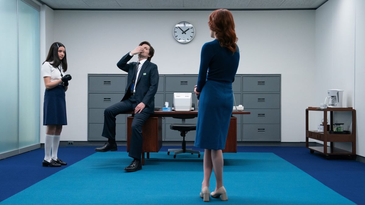 Mark S. sits on a desk holding his nose. Next to him, Miss Huang puts on black gloves. Across the room, with her back to the camera, Helly R. stands cross-legged. Severance Season 2 Episode 6, "Attila."