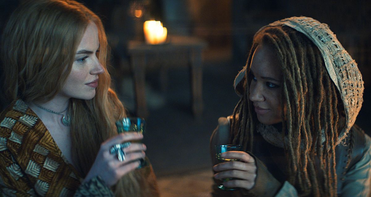A photo of Ceara Coveney as Elayne and Ayoola Smart as Aviendha in The Wheel of Time Season 3. They each hold glasses while sitting on the floor and gazing lovingly into each other's eyes.