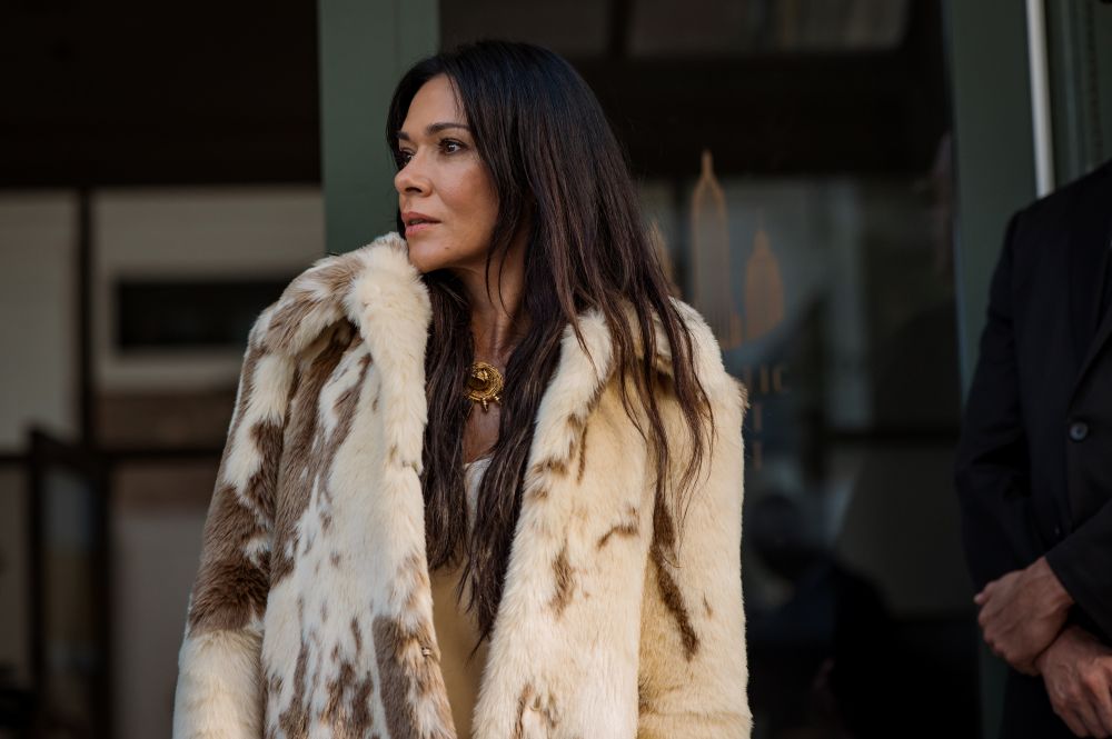 Lottie, a white woman, wears a white fur coat with brown spots. She stands outside an apartment complex in New York City during the day.