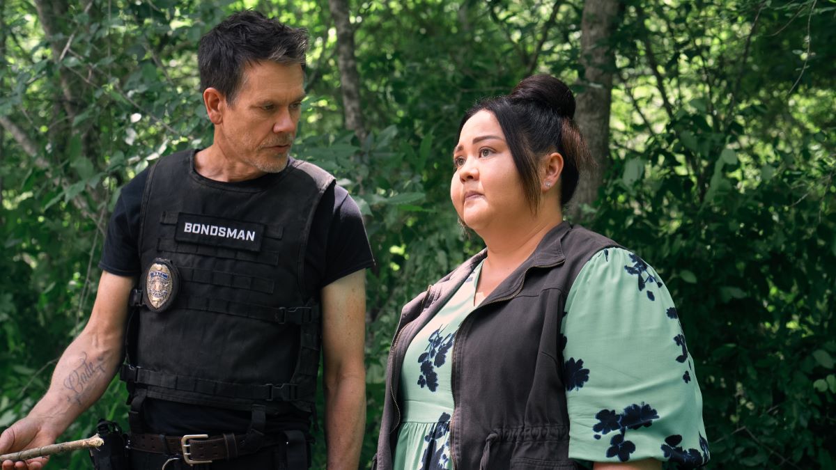 Hub and Midge confer out in the woods. He wears a bulletproof vest over a black t-shirt. She has on a light vest over a green floral print top. The Bondsman 108