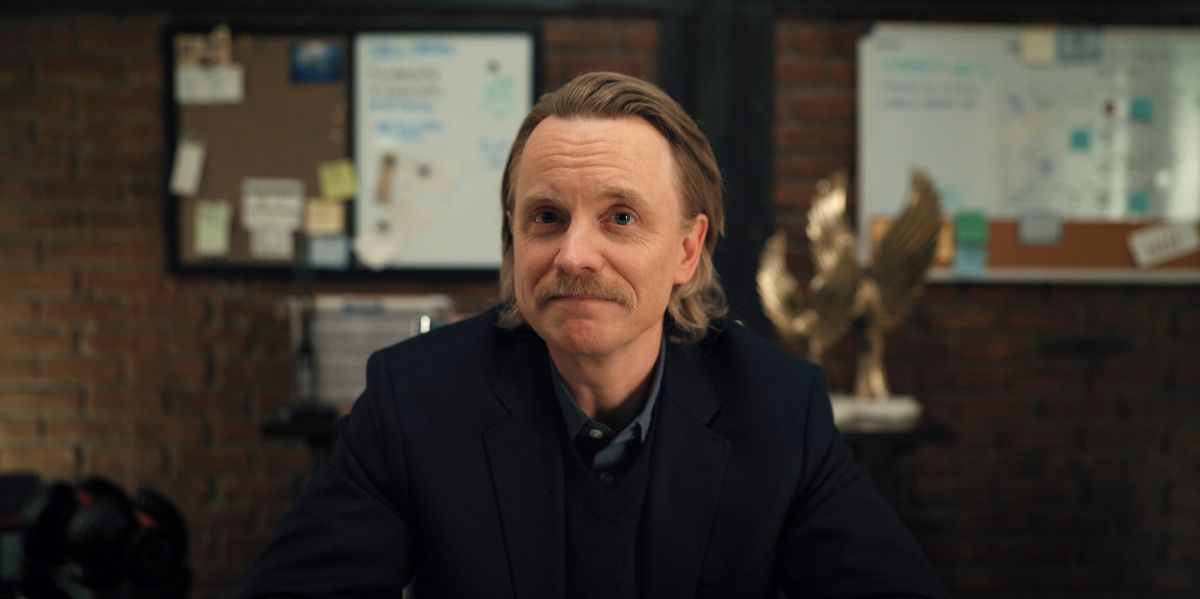 David, a middle-aged white man with blonde hair and a mustache, sits at his desk in his office at a video game company while smiling with his mouth closed.