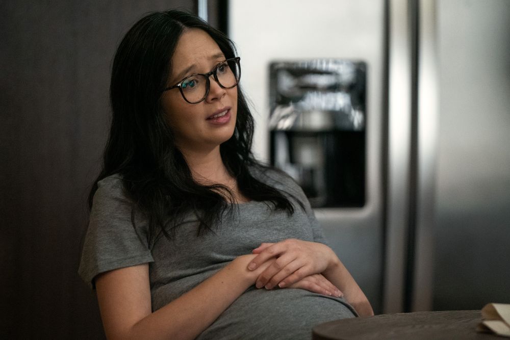 Poppy sits in a chair in her apartment kitchen with her hands resting on her pregnant stomach. She looks concerned.