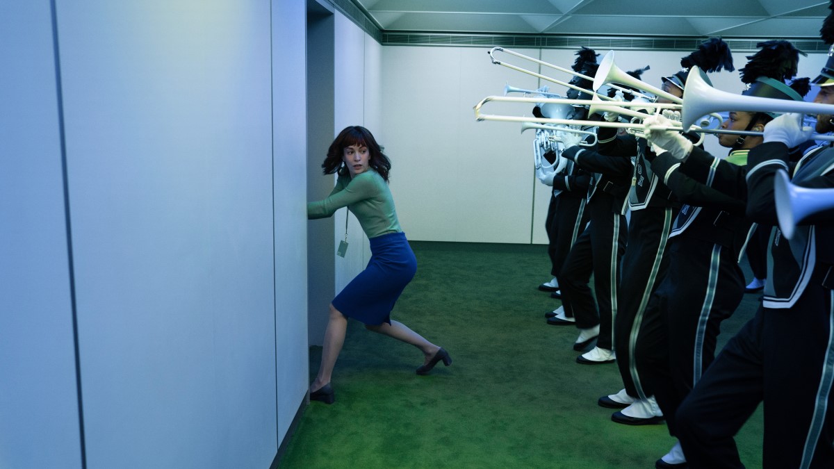 Helly R. holds a door closed while a marching band plays behind her in Severance Season 2.