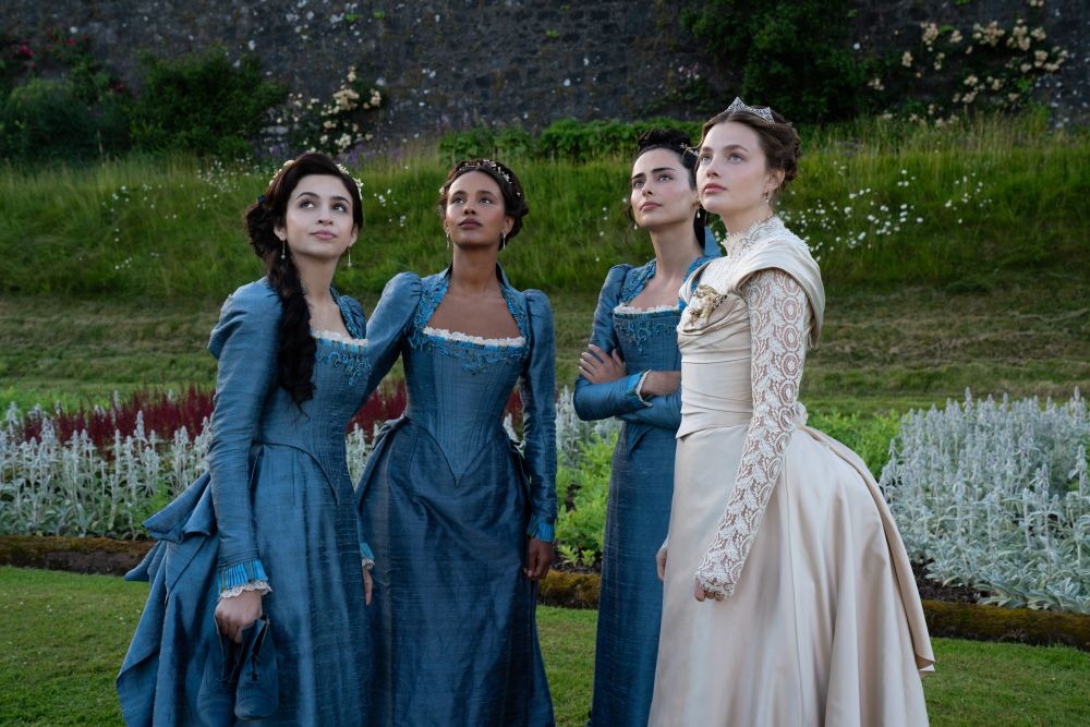 Mabel Elmsworth, Conchita Closson, Lizzy Elmsworth and Nan St. George stand outside on an overcast day in the countryside. Mabel, Conchita and Lizzy wear blue bridesmaids dresses while Nan wears a white bridal gown in The Buccaneers Season 2.