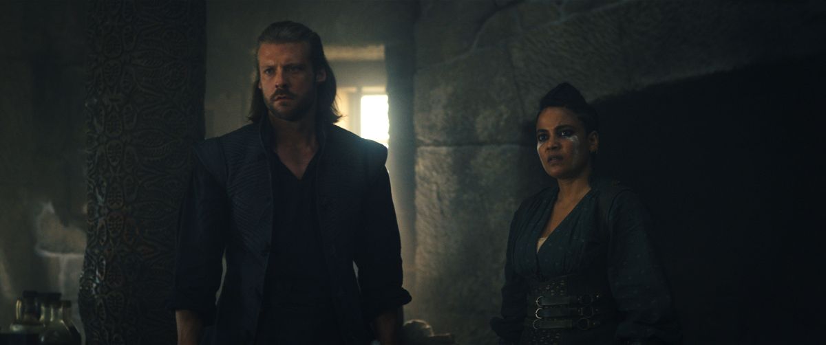 Taylor Napier as Maksim and Priyanka Bose as Alanna Sedai stand inside an inn in Emond's Field in the Two Rivers. They look at something out of frame with focus.