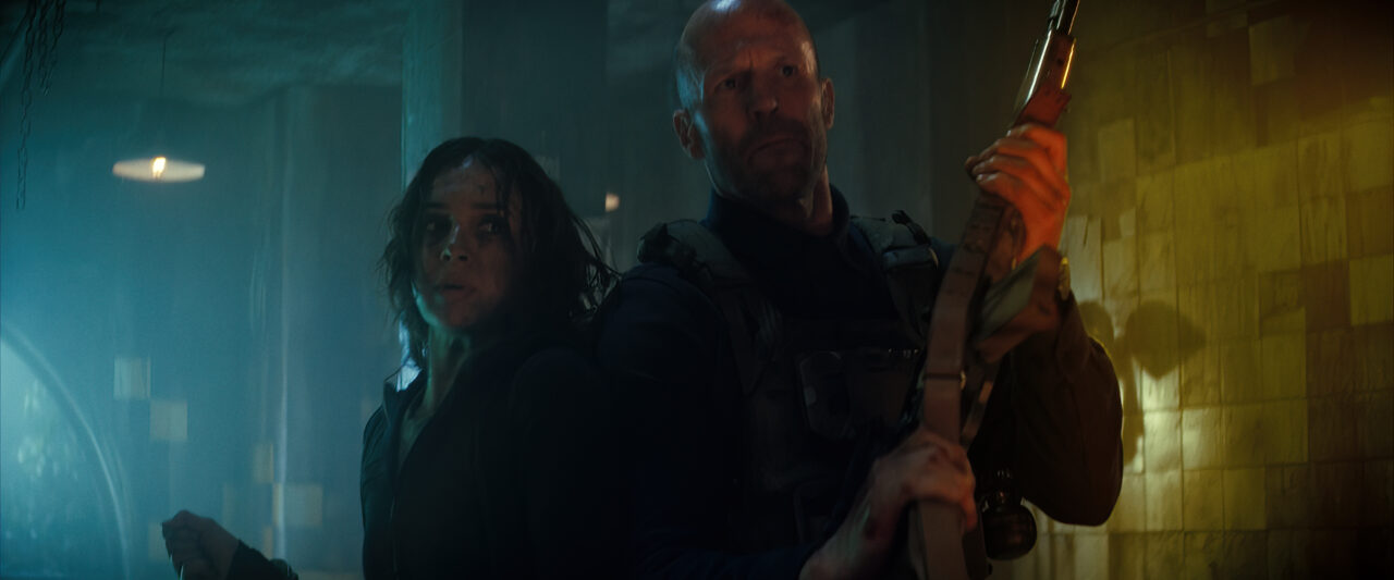 Arianna Rivas and Jason Statham stand back to back in a dark room.