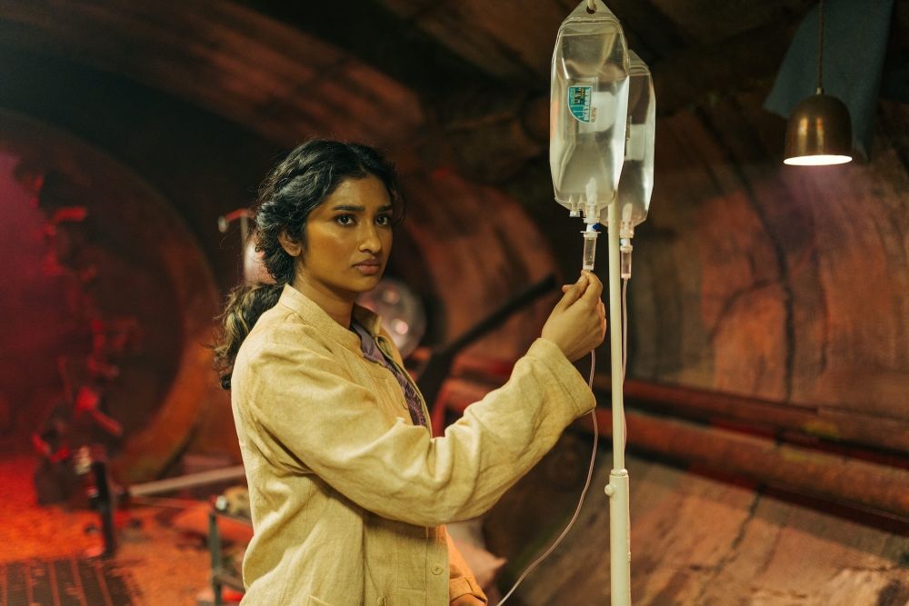 Belinda Chandra adjusts an IV stand while standing in an underground facility on a faraway planet on Doctor Who Season 2 Episode 1, "The Robot Revolution."