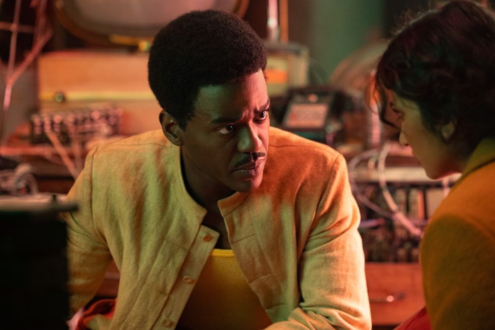 The Doctor wears a tan linen button-down top with matching linen pants. He talks to Belinda Chandra with a somber expression on his face while kneeling in an underground facility on a faraway planet.