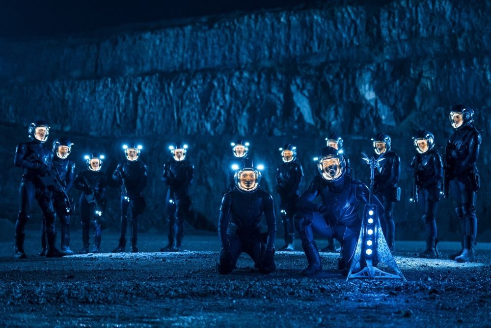 Belinda and the Doctor wear dark blue and black spacesuits while kneeling beside the Vindicator on an alien planet's surface. Troopers, also in the same gear, stand behind them on Doctor Who Season 2 Episode 3, "The Well."